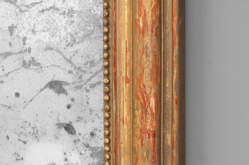 A Louis Philippe style wall mirror featuring a rounded rectangular giltwood frame with visible red bole ground, beaded inner trim, and heavily foxed glass. View V