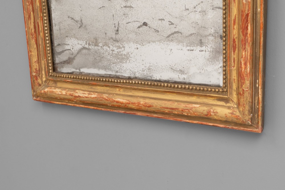 A Louis Philippe style wall mirror featuring a rounded rectangular giltwood frame with visible red bole ground, beaded inner trim, and heavily foxed glass. View VI