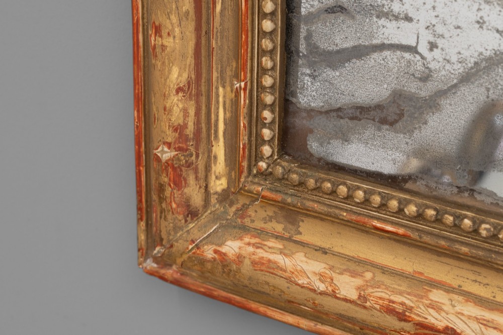 A Louis Philippe style wall mirror featuring a rounded rectangular giltwood frame with visible red bole ground, beaded inner trim, and heavily foxed glass. View VII