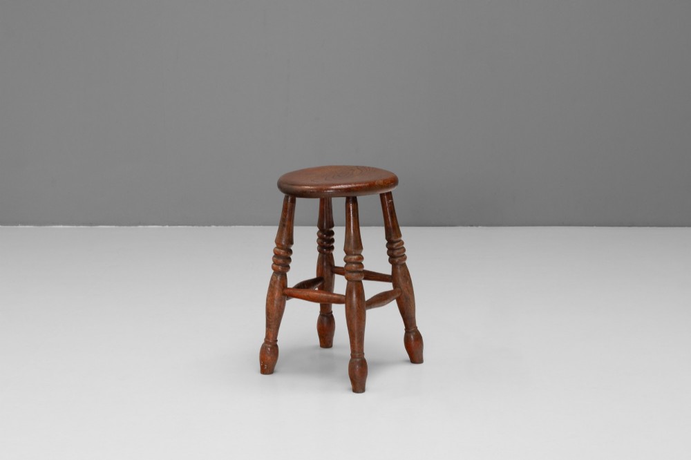 A dark brown wooden stool featuring a round seat, four lathe-turned legs, and lower connecting stretchers. View II