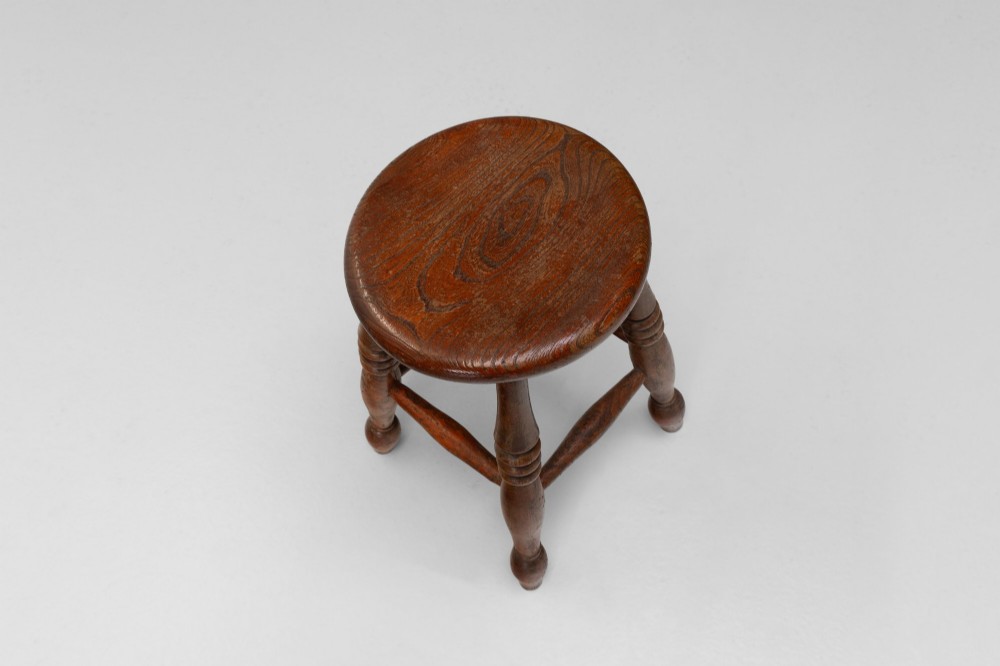 A dark brown wooden stool featuring a round seat, four lathe-turned legs, and lower connecting stretchers. View III