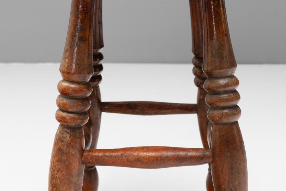 A dark brown wooden stool featuring a round seat, four lathe-turned legs, and lower connecting stretchers. View V