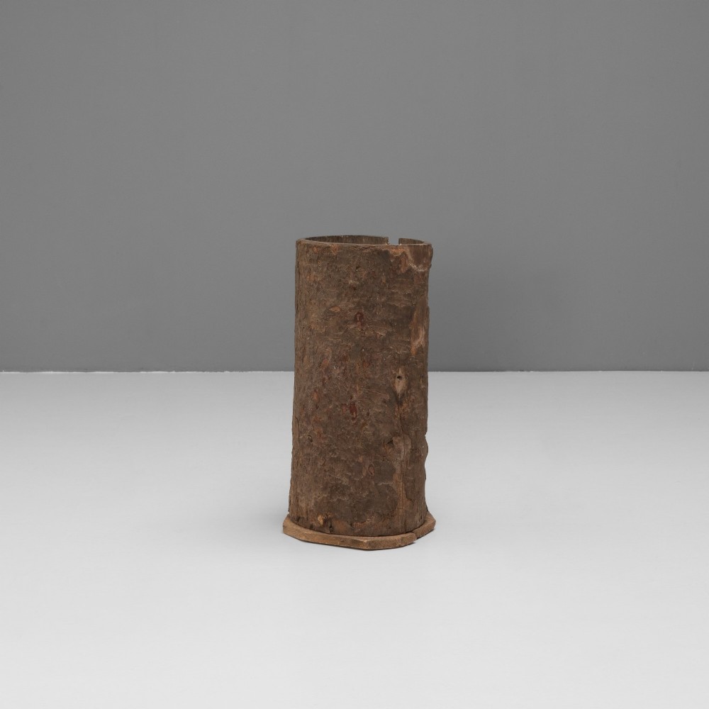 A tall cylindrical umbrella stand constructed from a hollowed tree trunk with original dark brown bark set on a circular wooden base.