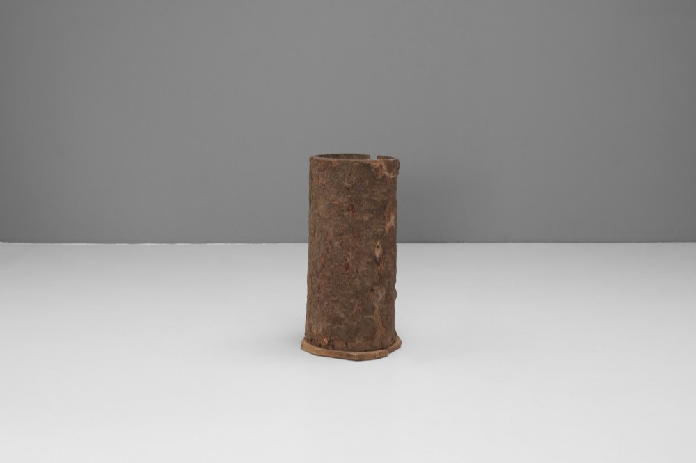 A tall cylindrical umbrella stand constructed from a hollowed tree trunk with original dark brown bark set on a circular wooden base. View II