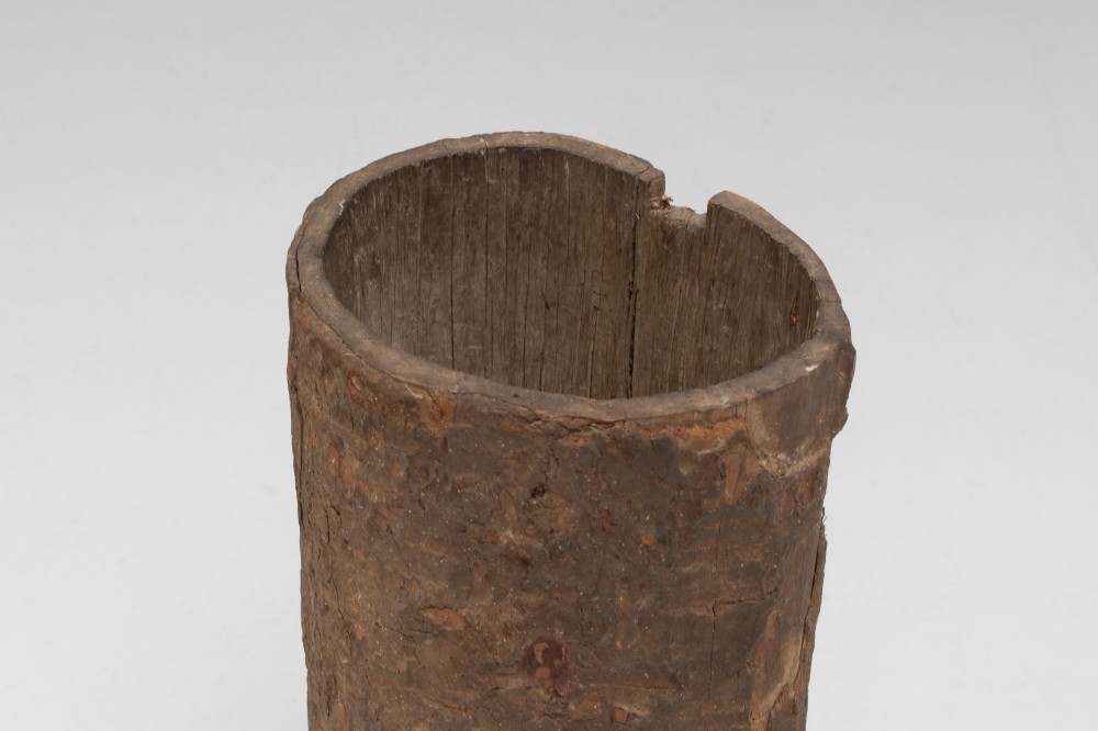 A tall cylindrical umbrella stand constructed from a hollowed tree trunk with original dark brown bark set on a circular wooden base. View III