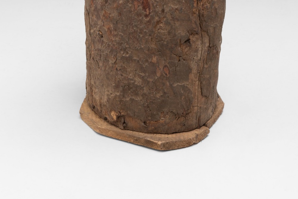 A tall cylindrical umbrella stand constructed from a hollowed tree trunk with original dark brown bark set on a circular wooden base. View IV