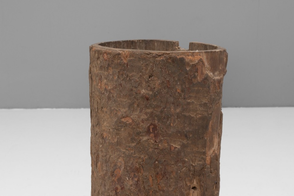 A tall cylindrical umbrella stand constructed from a hollowed tree trunk with original dark brown bark set on a circular wooden base. View V