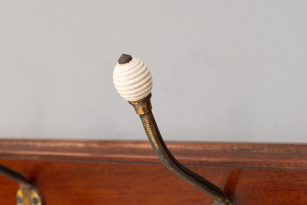 A Victorian wall-mounted mahogany coat rack featuring four brass hooks with white ceramic knobs set against a rectangular wooden backboard. View VI