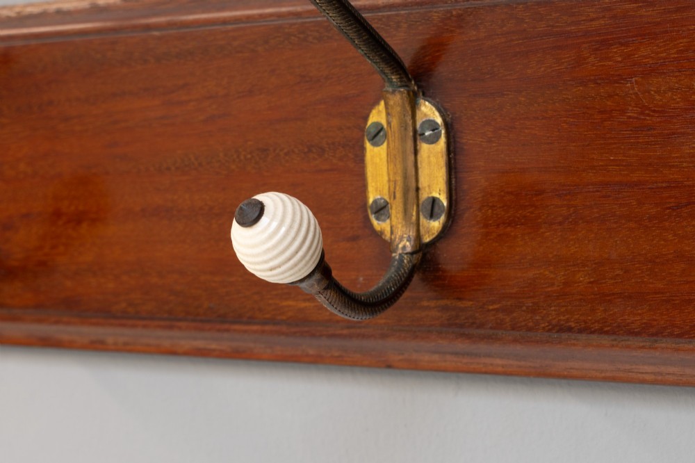 A Victorian wall-mounted mahogany coat rack featuring four brass hooks with white ceramic knobs set against a rectangular wooden backboard. View VII