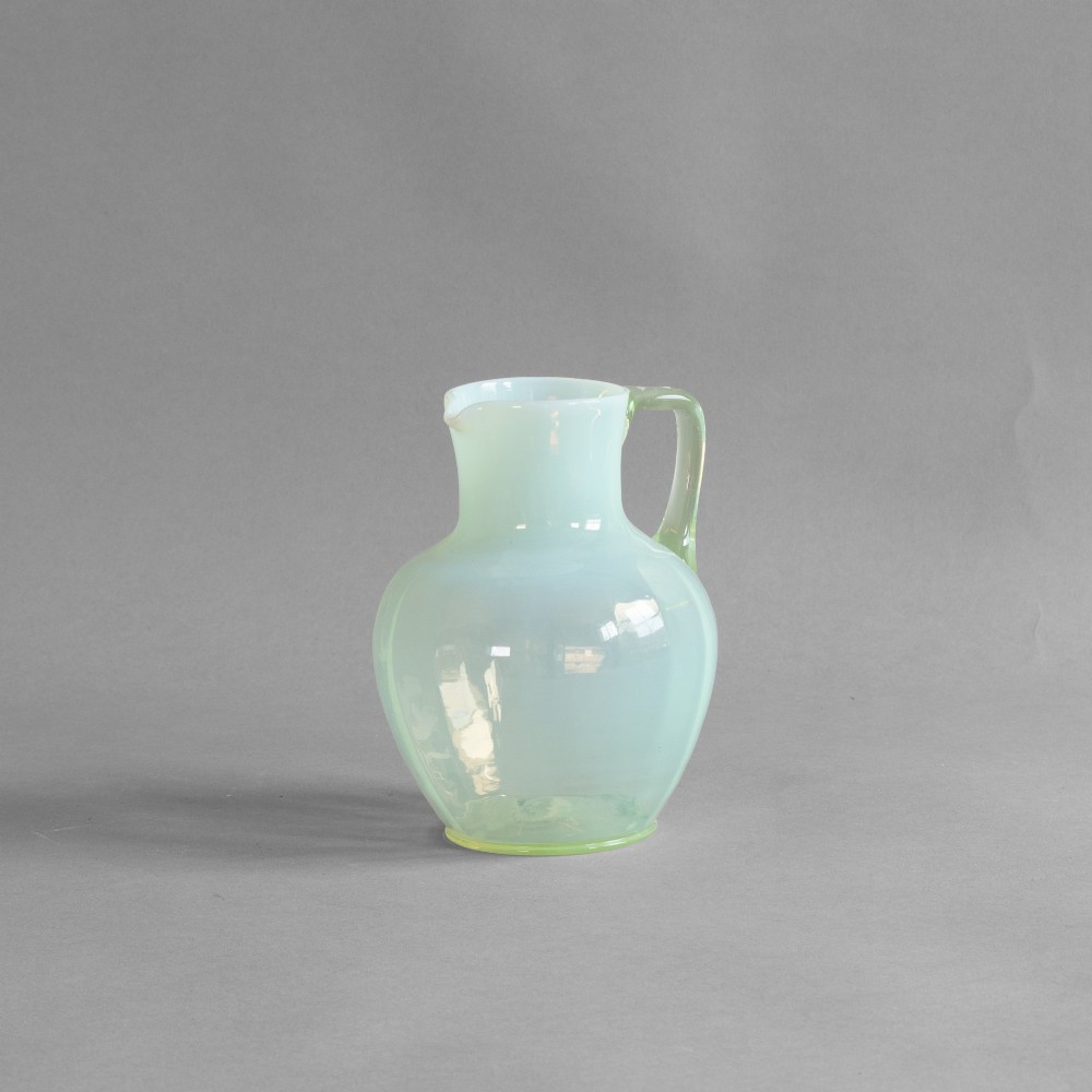 A pale green opalescent glass jug featuring a round bulbous body, flared neck, pinched spout, and an applied loop handle.