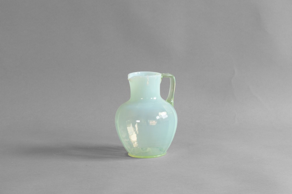 A pale green opalescent glass jug featuring a round bulbous body, flared neck, pinched spout, and an applied loop handle. View II
