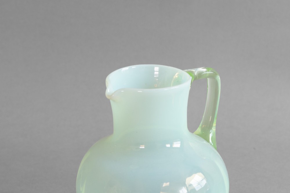 A pale green opalescent glass jug featuring a round bulbous body, flared neck, pinched spout, and an applied loop handle. View III