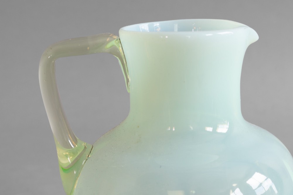 A pale green opalescent glass jug featuring a round bulbous body, flared neck, pinched spout, and an applied loop handle. View IV