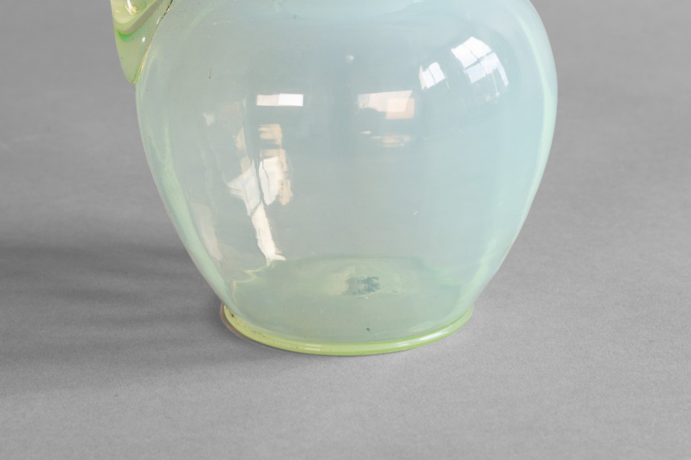 A pale green opalescent glass jug featuring a round bulbous body, flared neck, pinched spout, and an applied loop handle. View V