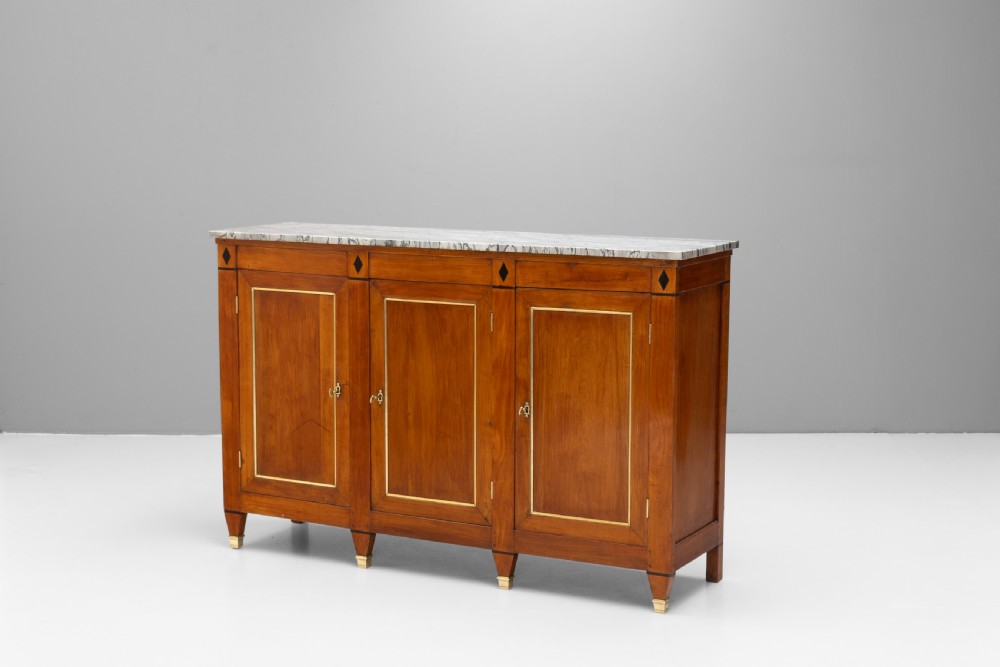 A three-door warm brown wooden sideboard featuring brass trim framing on panelled doors, dark diamond corner inlays, tapered legs, and a grey-veined white marble slab top.