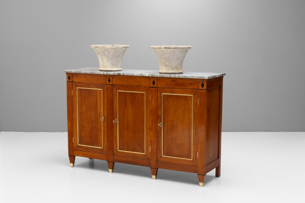 A three-door warm brown wooden sideboard featuring brass trim framing on panelled doors, dark diamond corner inlays, tapered legs, and a grey-veined white marble slab top. View II