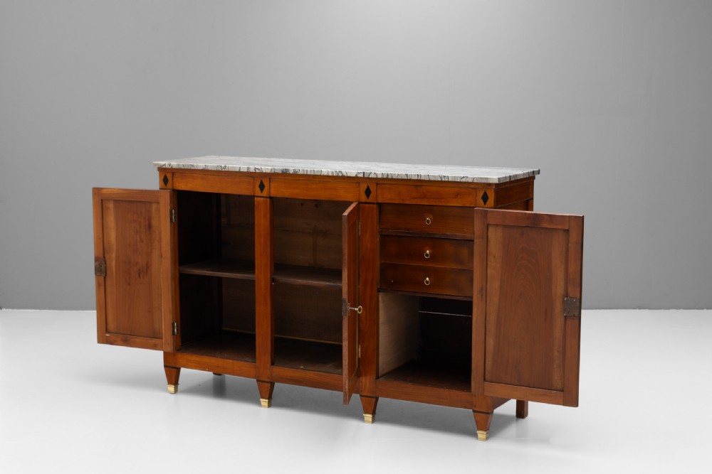 A three-door warm brown wooden sideboard featuring brass trim framing on panelled doors, dark diamond corner inlays, tapered legs, and a grey-veined white marble slab top. View III