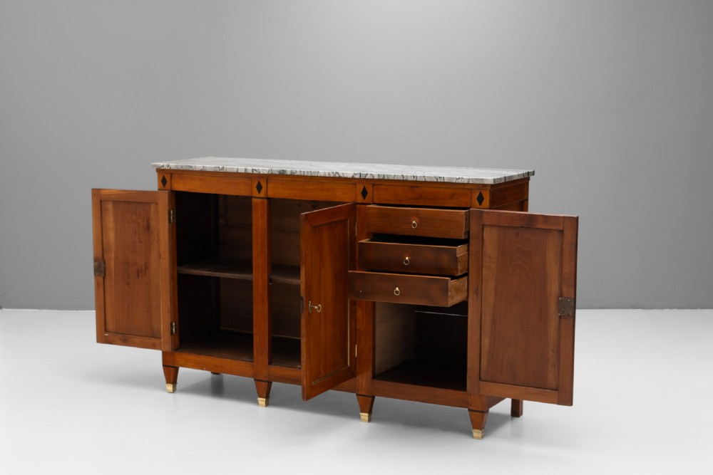 A three-door warm brown wooden sideboard featuring brass trim framing on panelled doors, dark diamond corner inlays, tapered legs, and a grey-veined white marble slab top. View IV