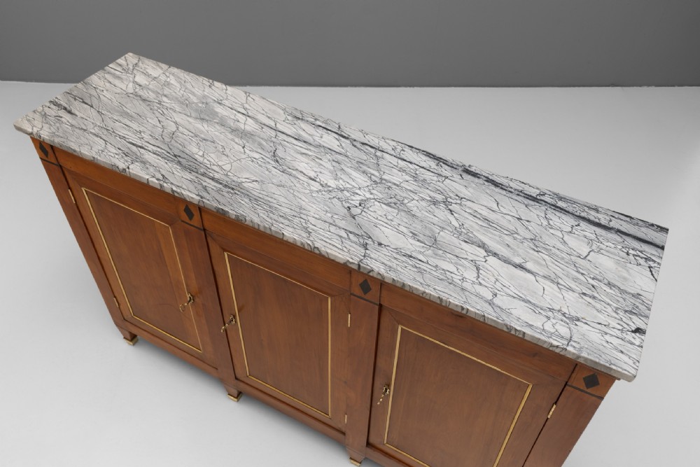 A three-door warm brown wooden sideboard featuring brass trim framing on panelled doors, dark diamond corner inlays, tapered legs, and a grey-veined white marble slab top. View V