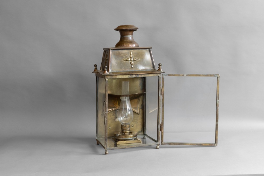 An antique glazed brass and copper lantern housing an internal glass oil lamp chimney and burner against a polished backplate. View III