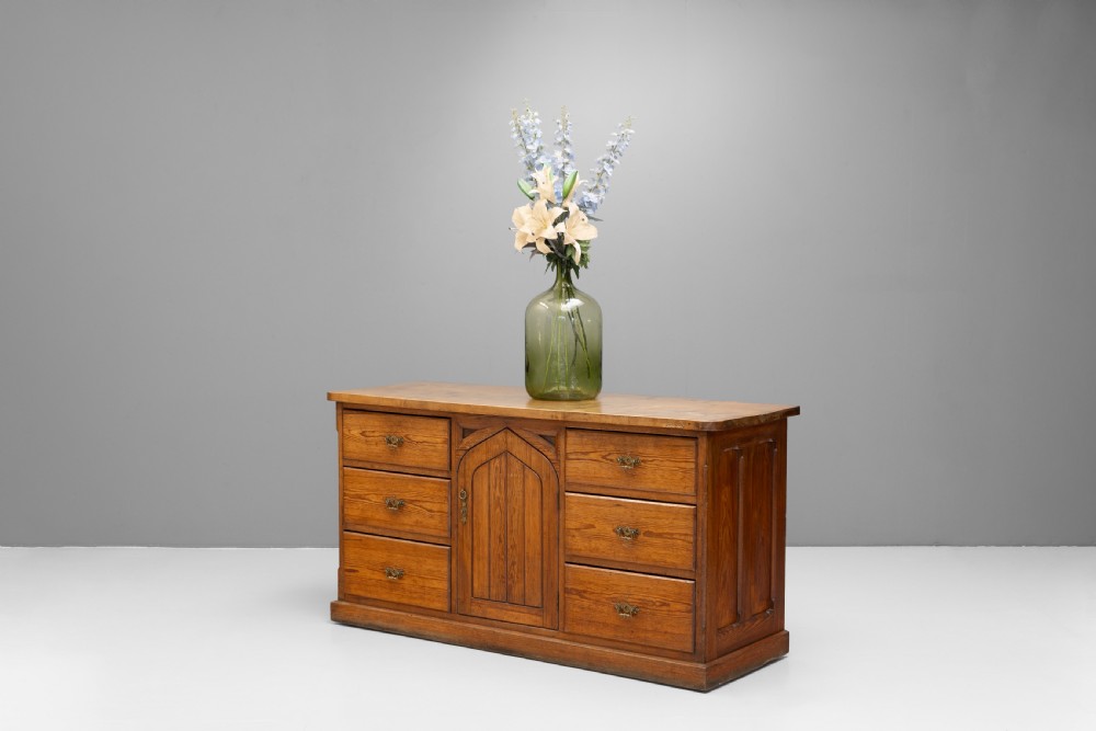 A warm brown pine sideboard featuring a central arched cupboard door flanked by six drawers with brass handles on a solid plinth base.