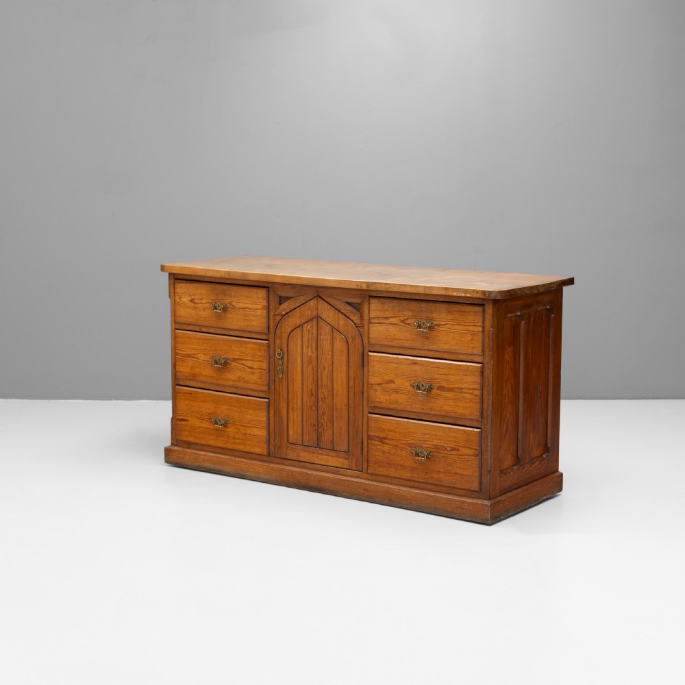 A warm brown pine sideboard featuring a central arched cupboard door flanked by six drawers with brass handles on a solid plinth base. View II