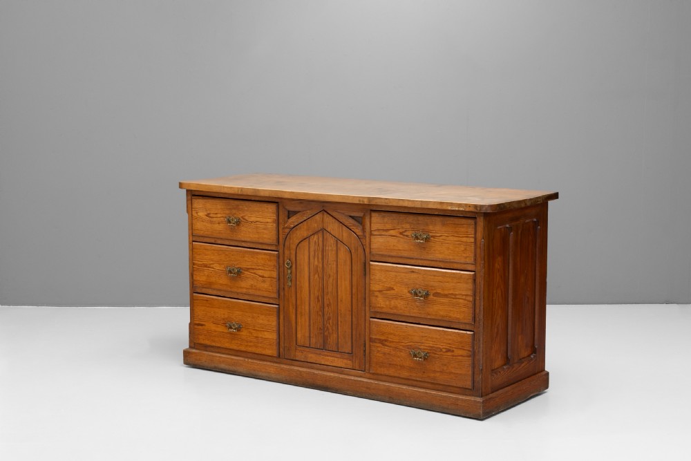 A warm brown pine sideboard featuring a central arched cupboard door flanked by six drawers with brass handles on a solid plinth base. View III
