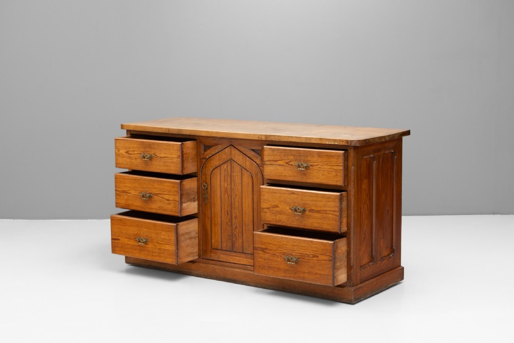 A warm brown pine sideboard featuring a central arched cupboard door flanked by six drawers with brass handles on a solid plinth base. View V