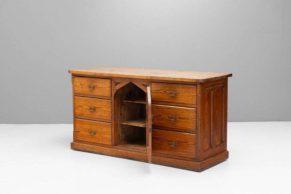 A warm brown pine sideboard featuring a central arched cupboard door flanked by six drawers with brass handles on a solid plinth base. View VI