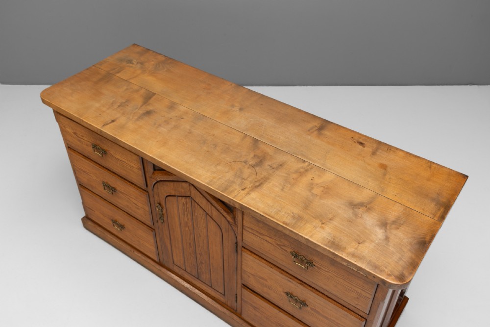A warm brown pine sideboard featuring a central arched cupboard door flanked by six drawers with brass handles on a solid plinth base. View VII