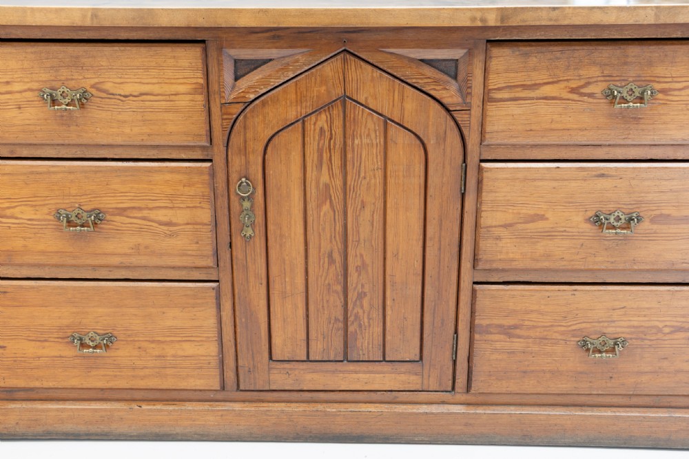 A warm brown pine sideboard featuring a central arched cupboard door flanked by six drawers with brass handles on a solid plinth base. View X