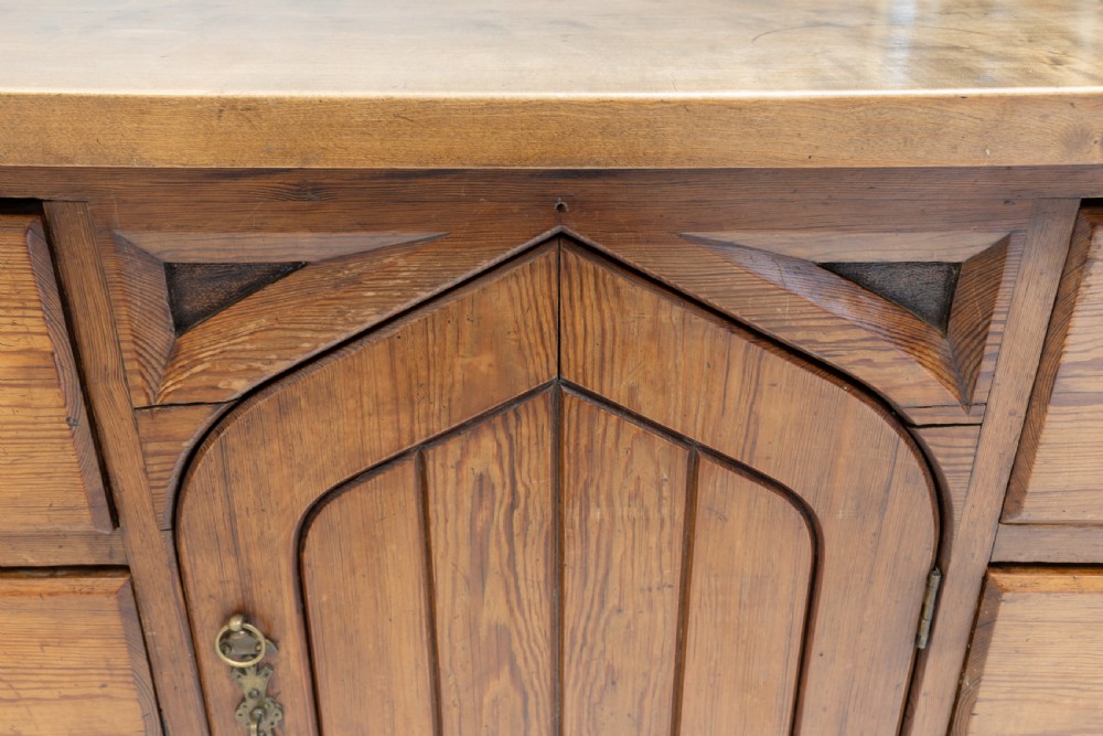 A warm brown pine sideboard featuring a central arched cupboard door flanked by six drawers with brass handles on a solid plinth base. View XI