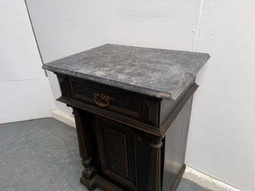 fig 1. A dark painted antique wooden bedside cabinet featuring a grey marble top, fluted architectural columns, a carved upper drawer with a brass pull, and a single cupboard door.