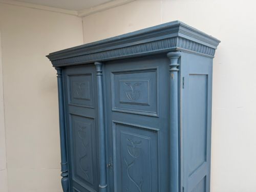 fig 1. A blue painted double-door wooden hall wardrobe featuring carved floral motifs on the panels, flanking turned columns, and a lower base drawer.