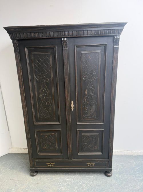 fig 1. A tall black painted timber hall wardrobe featuring carved floral door panels, a fluted cornice, a bottom drawer, and brass handles.