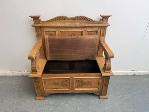 fig 1. A light oak Victorian box settle featuring carved floral and geometric panels, curved armrests, and a lift-up storage seat.