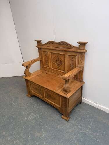 A light oak Victorian box settle featuring carved floral and geometric panels, curved armrests, and a lift-up storage seat.