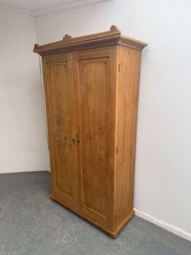 An Arts and Crafts waxed pine two-door wardrobe featuring shallow carved floral door panels and a carved top cresting rail.