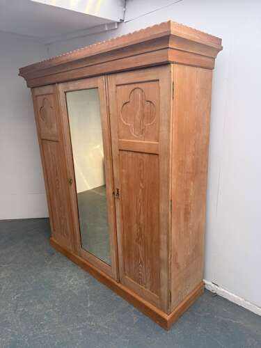 A three-door Arts and Crafts ash wardrobe featuring a central mirrored door flanked by panelled doors decorated with carved quatrefoil motifs.