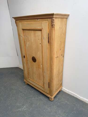 A tall Victorian stripped pine cupboard with a single panelled door, carved accents near the top corners, and turned bun feet.
