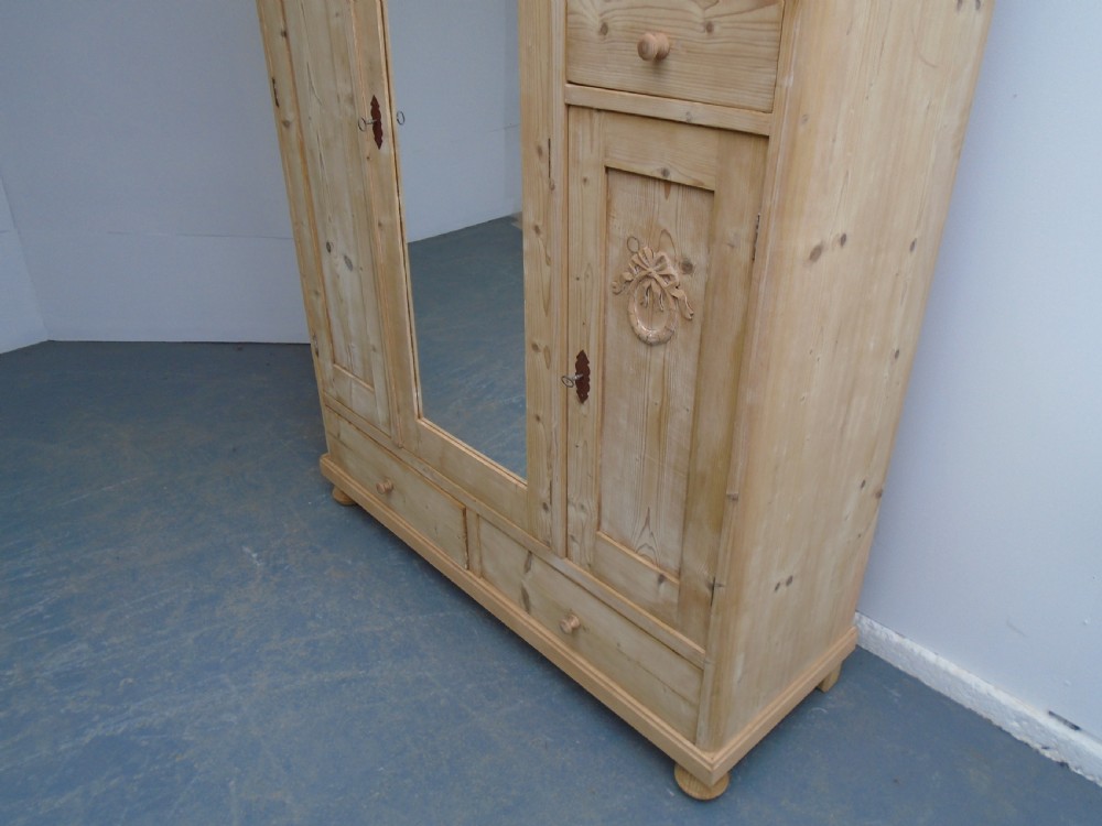 A large Victorian stripped pine wardrobe featuring a central mirrored door, carved ribbon relief motifs, an open alcove, drawers, and turned bun feet. View III