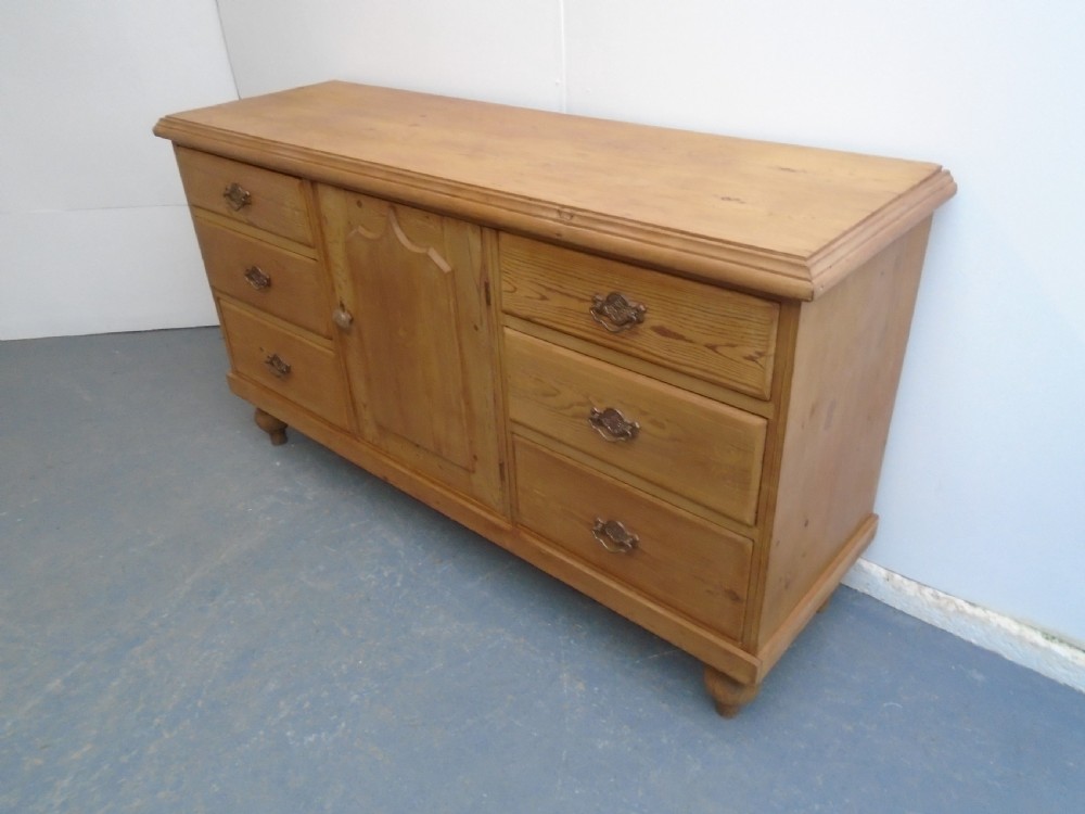 A Victorian waxed pine dresser base featuring six drawers flanking a central panelled cupboard door, raised on turned bun feet with metal drop handles.