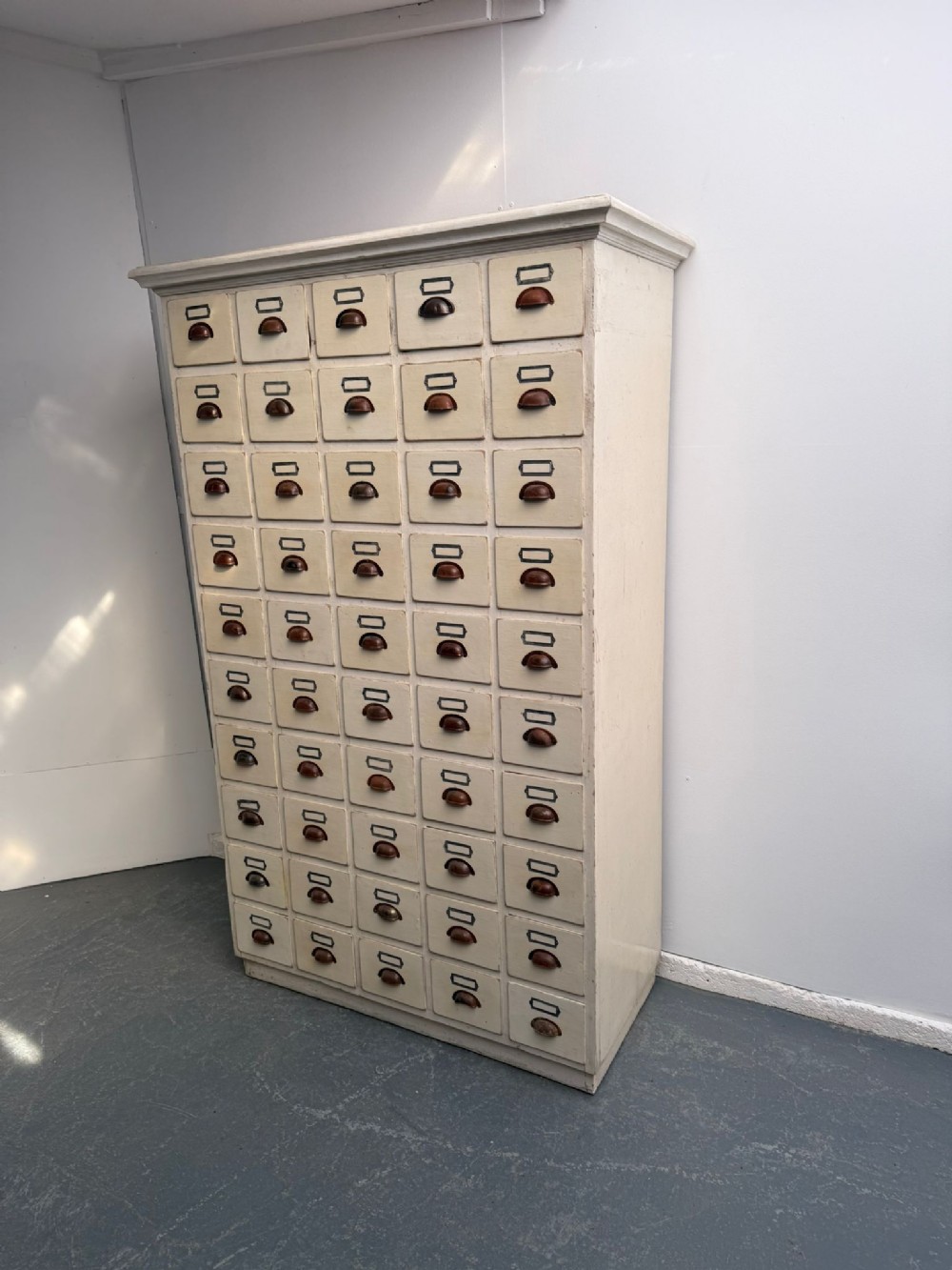 A tall off-white painted pine cabinet featuring fifty individual drawers with dark metal shell handles and label holders.