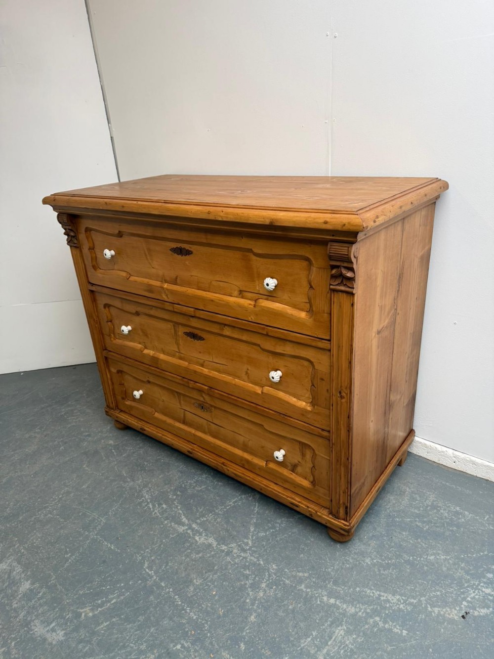 A waxed pine chest of drawers featuring three wide drawers with shaped panel details and white ceramic handles.