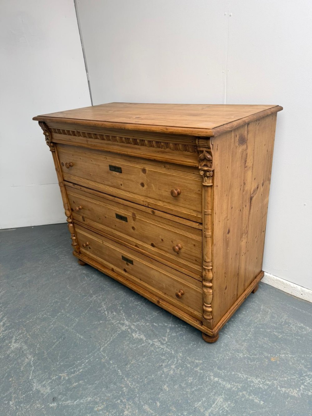A three-drawer waxed pine chest featuring side columns with carved capitals, turned wooden knobs, and inset brass keyhole escutcheons.