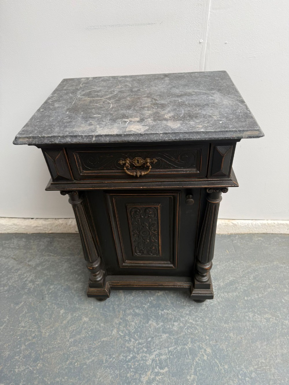 A dark painted antique wooden bedside cabinet featuring a grey marble top, fluted architectural columns, a carved upper drawer with a brass pull, and a single cupboard door.
