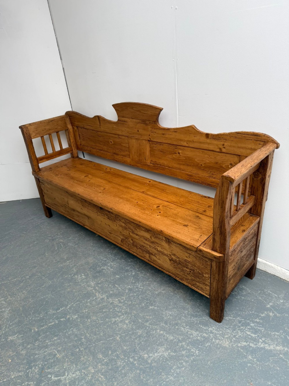 A long waxed pine box settle featuring a contoured crest backrest, slatted side arms, and an integrated storage seat base.