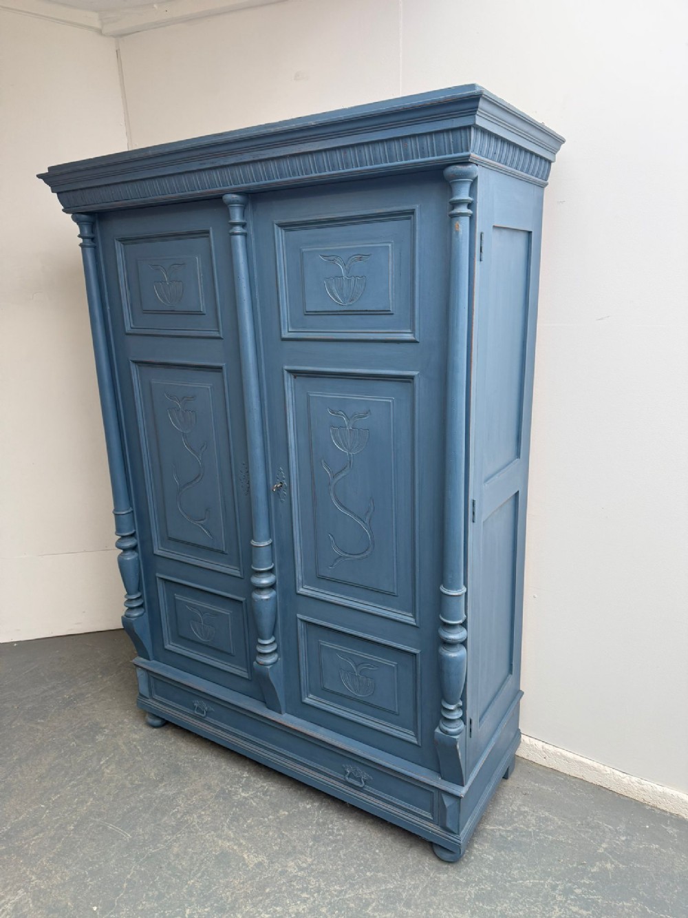 A blue painted double-door wooden hall wardrobe featuring carved floral motifs on the panels, flanking turned columns, and a lower base drawer.