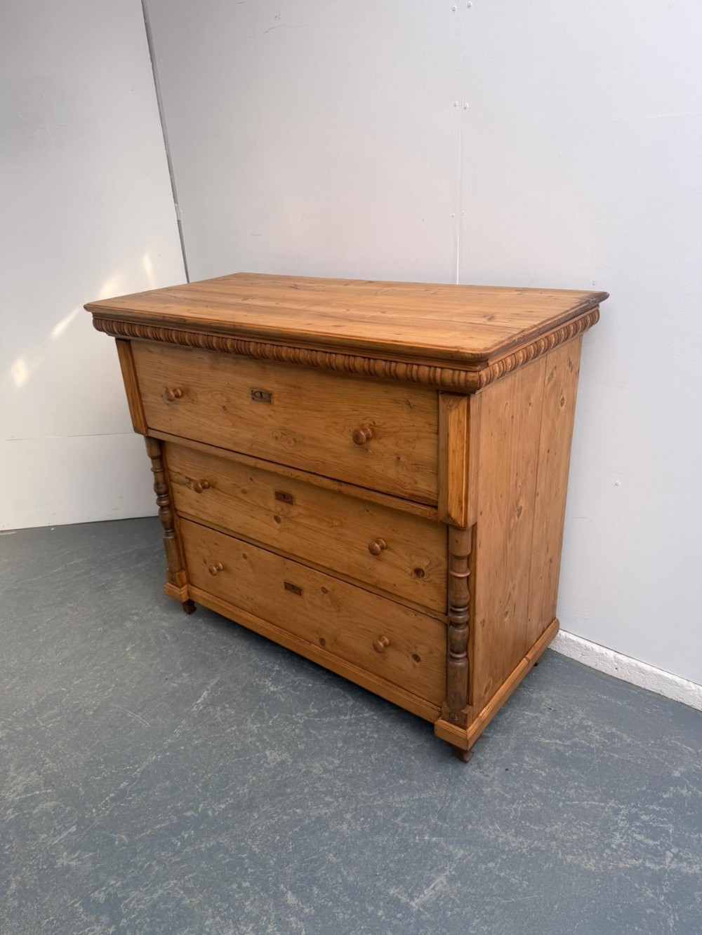 A waxed pine chest of drawers featuring three full-width drawers with turned wooden knobs, flanked by carved side columns and topped with carved trim.