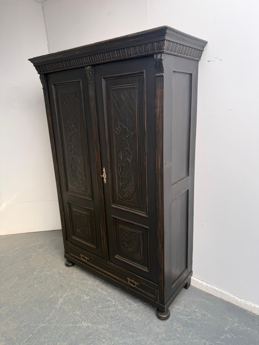 A tall black painted timber hall wardrobe featuring carved floral door panels, a fluted cornice, a bottom drawer, and brass handles.
